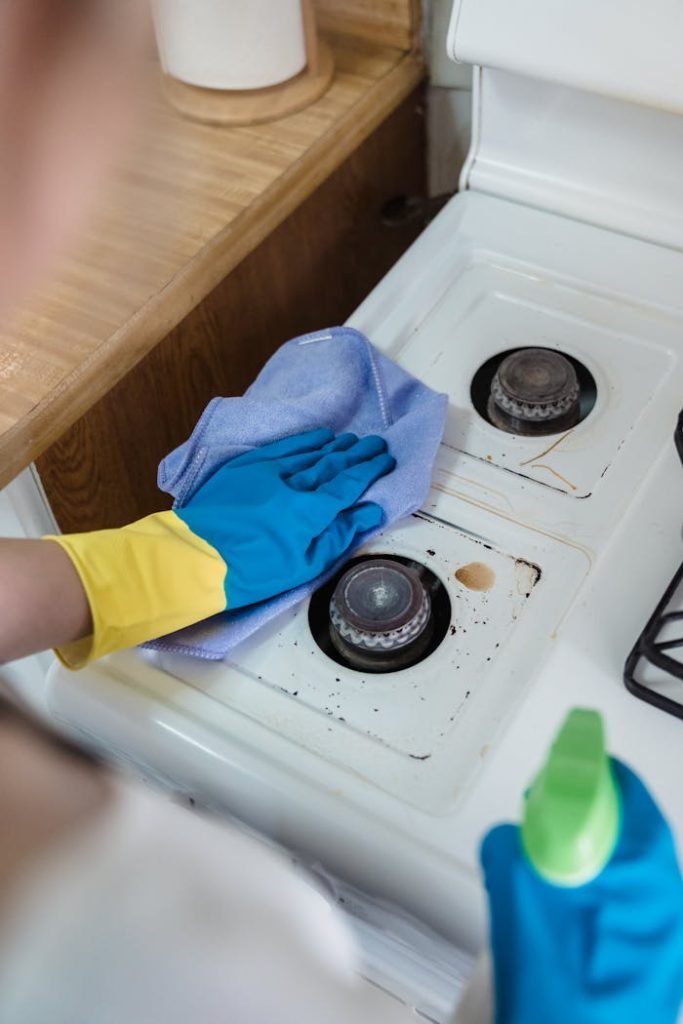 a-person-cleaning-a-gas-stove-9462302 A person wearing rubber gloves cleaning a dirty gas stove with a cloth and spray bottle.