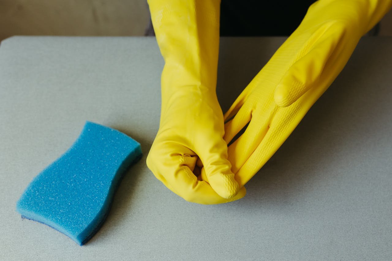 about-img Close-up of hands wearing yellow gloves next to blue sponge on gray surface.
